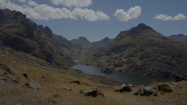 High-angle wide shot of a deep blue mountain lake nestled within a vast Andean canyon in Peru. Rugged rocky peaks, arid highland slopes, clear blue sky with clouds. Scenic daytime travel b-roll.