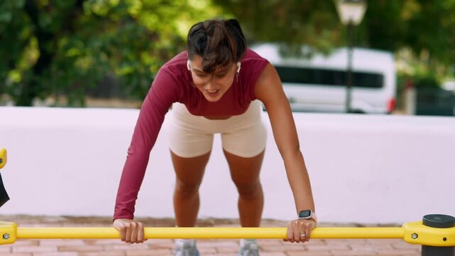 Young woman doing incline pushups on yellow parallel bars outdoors, low frontal angle focused on strength and controlled technique in city park daylight, energetic workout