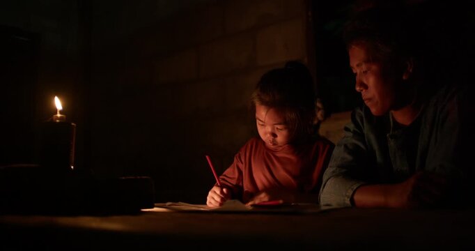 A girl colors in an activity workbook by oil lamp light at night, her mother sitting close beside her in a dim rural Thai home.
