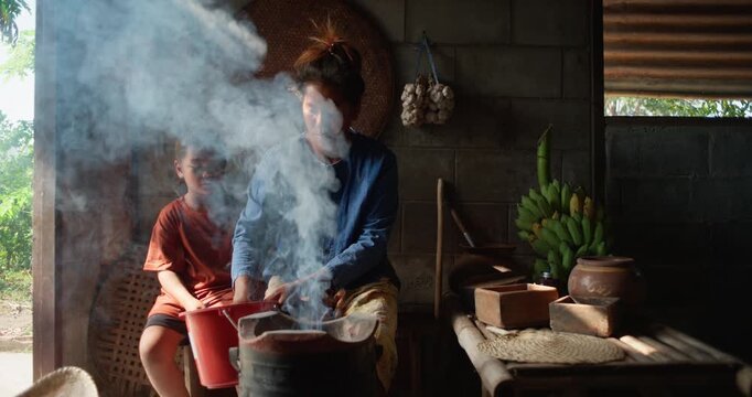 A Thai woman blows a charcoal stove to life, smoke curling in sunlight while her daughter watches beside her in a rural kitchen.