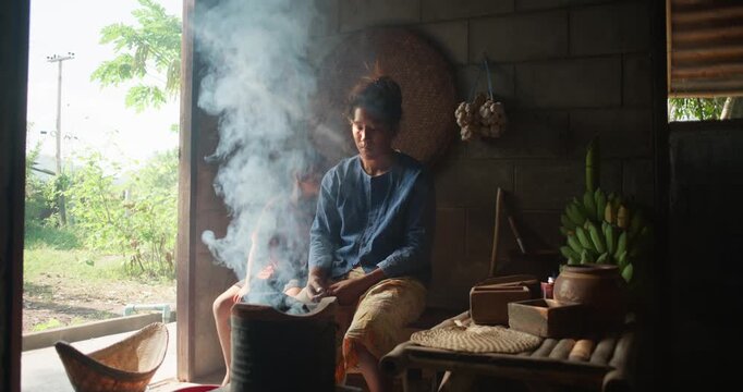 A Thai woman blows a charcoal stove to life, smoke curling in sunlight while her daughter watches beside her in a rural kitchen.