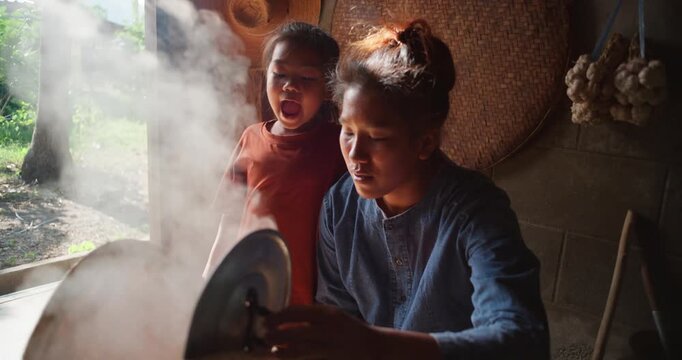Steam rises from a woven bamboo basket as sticky rice steams over a clay pot, with a daughter sitting nearby in a rural Thai kitchen.