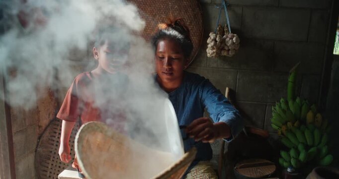 Steam rises from a woven bamboo basket as sticky rice steams over a clay pot, with a daughter sitting nearby in a rural Thai kitchen.