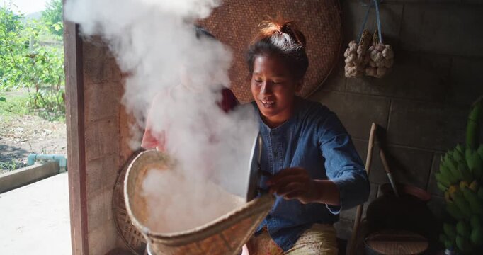 Steam rises from a woven bamboo basket as sticky rice steams over a clay pot, with a daughter sitting nearby in a rural Thai kitchen.