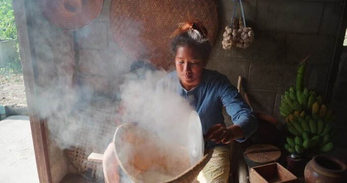 Steam rises from a woven bamboo basket as sticky rice steams over a clay pot, with a daughter sitting nearby in a rural Thai kitchen.