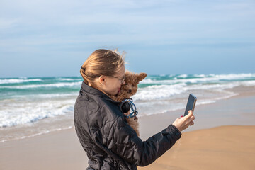 A young woman holds her little dog on arms while taking a selfie on a windy sandy beach coast. Autumn or winter oceanside travel recreation layout. Travel theme. © Natalia