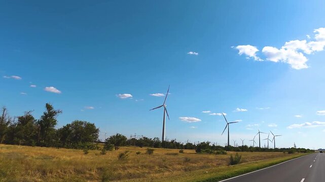 Wind turbines rotating in a vast agricultural field under a clear blue sky, with a paved road running alongside the landscape in a rural setting