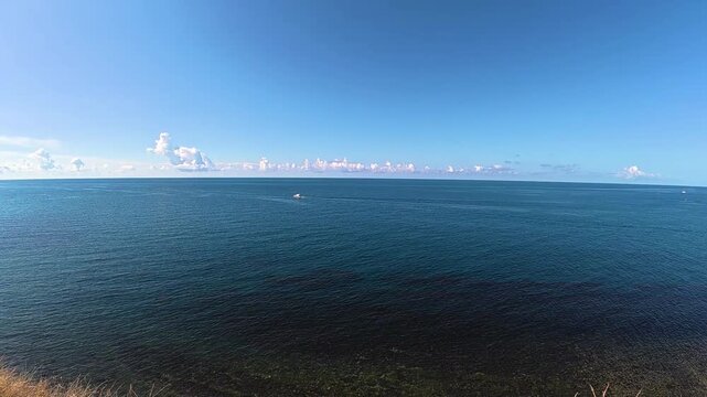 Scenic coastal view featuring a small boat navigating the calm blue waters under a clear sky with scattered clouds and a grassy shoreline in the foreground