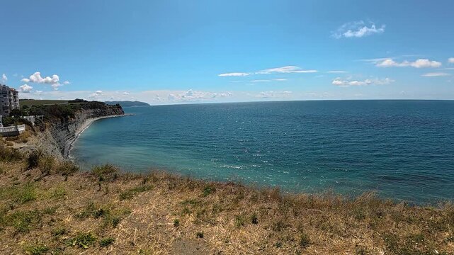Scenic coastal view featuring a small boat navigating the calm blue waters under a clear sky with scattered clouds and a grassy shoreline in the foreground