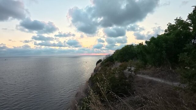 Coastal cliffside path overlooking calm sea beneath glowing sunset clouds and golden dry grass, tranquil natural landscape video