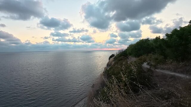 Coastal cliffside path overlooking calm sea beneath glowing sunset clouds and golden dry grass, tranquil natural landscape video