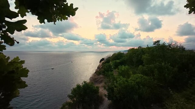 Calm sea stretches beside a rugged coastal cliff as leafy branches frame a narrow dirt path beneath a pastel sunset sky