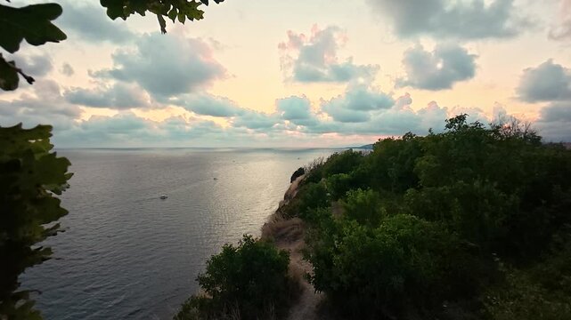 Coastal cliff overlooking calm sea beneath leafy branches and pastel sunset sky, with a narrow dirt path through dry grass and shrubs
