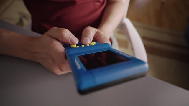 Hands playing vintage style handheld electronic game. Close-up hands of unrecognizable person presses yellow buttons on blue console, focused on entertainment nostalgia, closeup, slow motion.