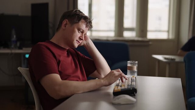 Tired young adult man feeling bored and uninspired at home office desk, props head on hand, procrastinating and struggling with creative block, burnout. Apathetic freelance worker, slow motion.
