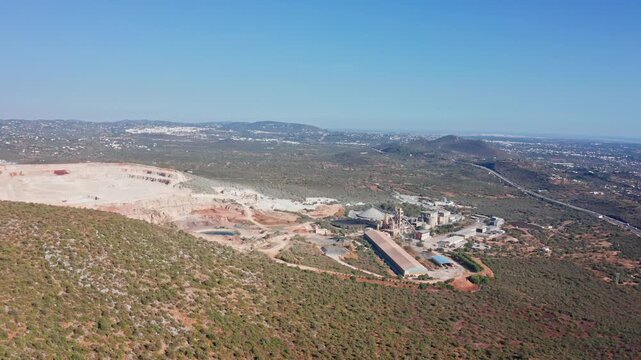 Aerial footage showing a vast open pit quarry and an adjacent industrial processing plant nestled in a hilly, green landscape under a clear blue sky, highlighting resource extraction and production