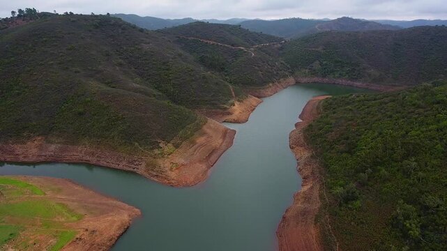 Aerial view of a winding river flowing through a dry, mountainous landscape, showing the effects of drought with low water levels and exposed, eroded banks under a cloudy sky