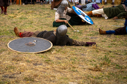 People wearing helmets and armor lie and sit on a grassy field beside shields during a medieval combat reenactment.