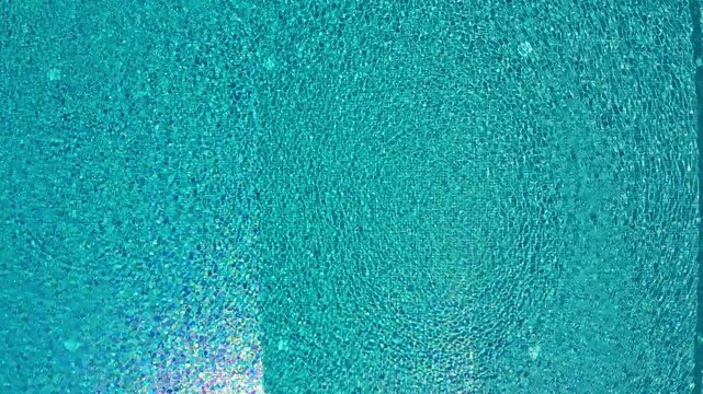 Top down aerial view slowly ascending from the turquoise water surface, revealing a modern rectangular swimming pool with steps and a stone deck, showcasing a luxurious summer vacation spot
