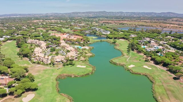 Aerial view flying over a prestigious golf course with a large green lake, luxury villas, and swimming pools in the beautiful resort town of vale do lobo, algarve, portugal