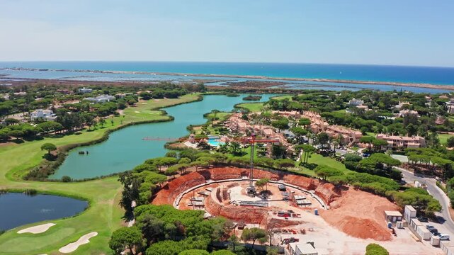 Stunning aerial perspective showing a large circular construction site for a new building next to a lush golf course, luxurious villas, a serene lake, and the beautiful blue ocean coast