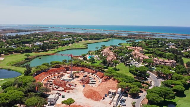 Aerial view of a large circular construction site for a new building in a luxury golf course resort near the ocean, showing excavation and foundational work with a crane in the center