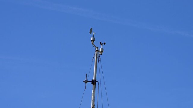 A marine cup anemometer and wind vane mounted atop a sailboat mast measure wind speed and direction against a clear blue sky.