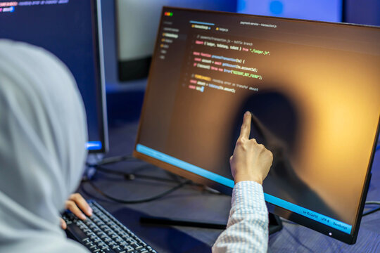 Female software engineer wearing a hijab pointing at computer screen with program code, debugging software and developing applications in a professional office. Technology, innovation concept.