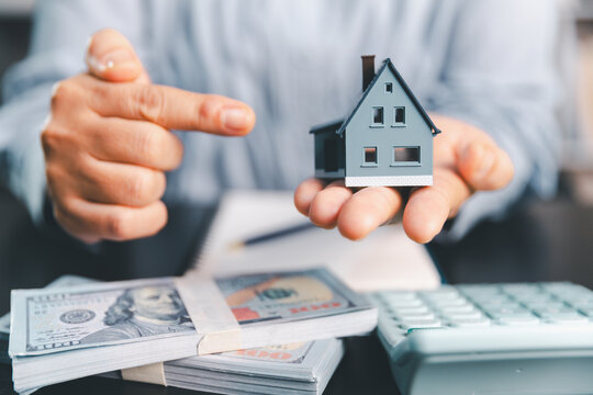 Person presents a model house above stacks of cash and a calculator, illustrating mortgage planning, home buying, property finance, loan budgeting, down payment goals