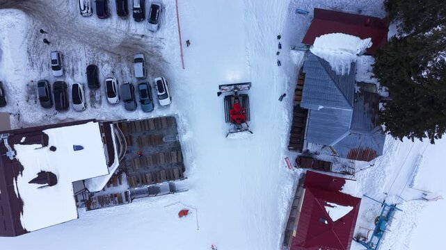 Red snowcat machine grooming snow at ski resort base, top down aerial view. A bright track vehicle stands on white ground near parked automobiles and wooden rooftops covered with fresh powder.
