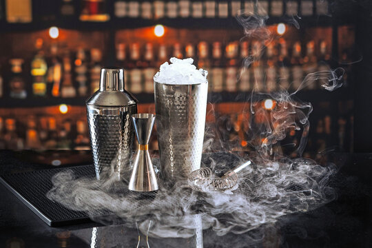 Bar tools with dry ice smoke and crushed ice. Shaker jigger strainer on black mat. Dark blurred bar background