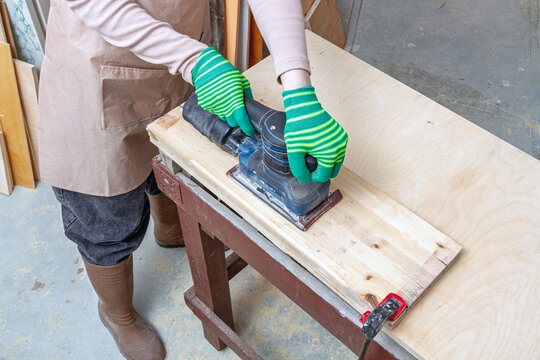 Worker using an orbital sander on a wooden board at a workbench. Woodworking and surface preparation. DIY furniture repair and renovation. Power tool operation for woodworking projects