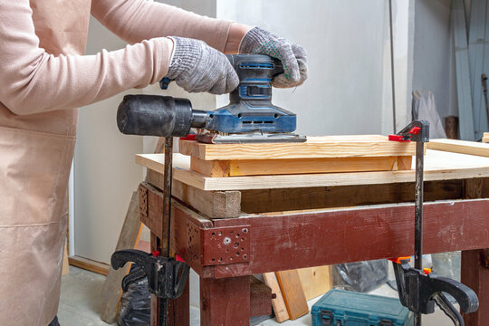 Person operating an orbital sander on a clamped wooden board at a workbench. Woodworking and furniture making. DIY workshop project. Power tool surface preparation and finishing