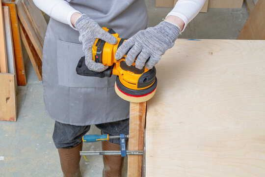 Worker using an orbital sander on a wooden board in a workshop. Woodworking and surface finishing. DIY furniture repair and renovation. Carpentry, sanding and tool use