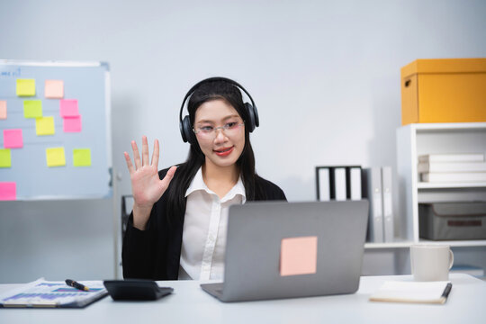 Young woman wearing headphones waving hand during video call on laptop in modern office with whiteboard and shelves