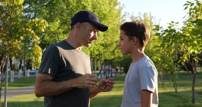 Father talks with teenage son in sunny park. Man removes glasses and explains advice during outdoor conversation. Teen listens with trust and curiosity. Family communication creates warm summer mood.