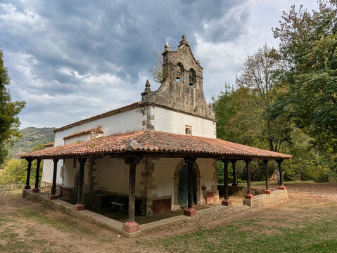 Rustic hermitage with wooden-columned porch, terracotta tile roof and stone bell gable under a stormy sky, Sanctuary of Our Lady of Miravalles, Aller, Asturias, Spain