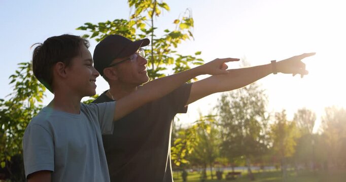 Dad and son point toward nature in park at sunset. Family bonding and outdoor discovery create warm summer evening mood. Guidance, curiosity, and joyful learning inspire active lifestyle memories