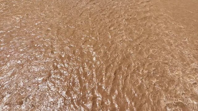 Aerial view shows fast moving muddy floodwater with turbulent waves, representing extreme weather, climate change, rain bomb rainfall, soil erosion and increasing flood risk.
