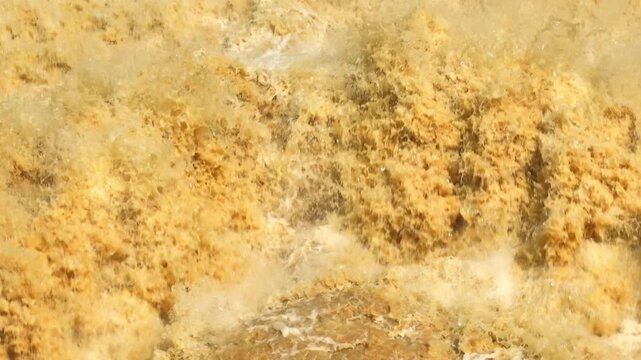 Aerial view captures powerful muddy floodwater released from a dam, showing violent turbulence, sediment rich currents, high volume discharge, flood control, and reservoir management.
