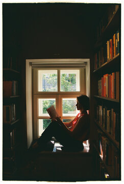 Person reads a book sitting in a window seat surrounded by bookshelves