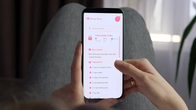 Woman opens a chocolate cake recipe on a smartphone and scrolls through ingredients, highlighting digital baking, home cooking, dessert preparation and modern recipe apps.