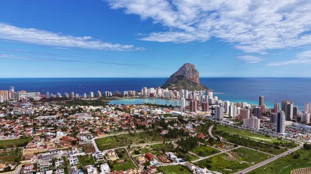 Aerial panoramic view of Calpe, Spain, featuring the iconic Penon de Ifach rock, turquoise Mediterranean Sea, salt lake, urban skyline, and residential neighborhoods under blue sky