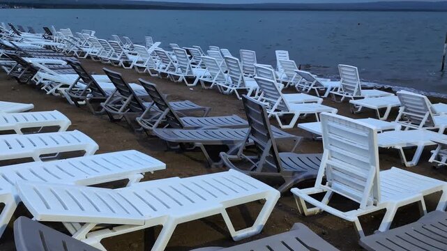 Rows of vacant white and brown lounge chairs line a sandy beach beside a calm but gray sea. Cloudy and stormy weather casts a moody atmosphere over the deserted shoreline, emphasizing the absence of.