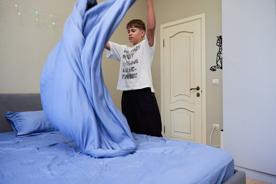 Teen boy making bed tossing blue blanket in bedroom