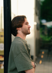 Side profile portrait of a young Caucasian man in a green t-shirt leaning against a black pole outdoors in a city street, looking away with crossed arms. © Vika