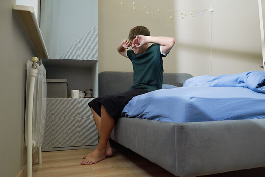 Sleepy teen boy rubbing eyes waking up on bed
