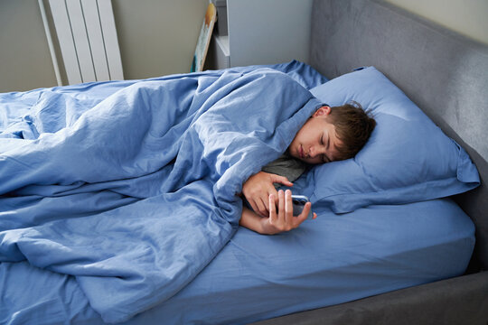Tired teen boy lying in bed holding smartphone falling asleep