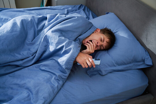 Sleepy teen boy yawning in bed holding smartphone