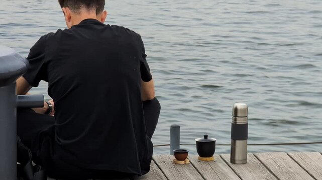 Man dressed in black clothes seated on a wooden dock by a body of water. Items including a tea pot, a small cup, and a metal thermos are placed on the dock beside him.